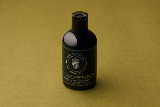 A black bottle of Crown Shaving Co. Aftershave Lotion sits on an olive green surface, its label facing forward and highlighting its reputation as a moisturizing balm ideal for sensitive skin.