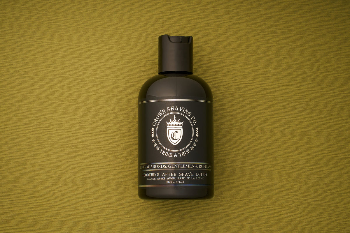 A black bottle of Crown Shaving Co. Aftershave Lotion stands upright on a green textured surface, displaying the product label and crest logo clearly—an ideal aftershave designed to soothe and moisturize sensitive skin.