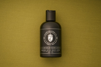 A black bottle of Crown Shaving Co. Aftershave Lotion stands upright on a green textured surface, displaying the product label and crest logo clearly—an ideal aftershave designed to soothe and moisturize sensitive skin.