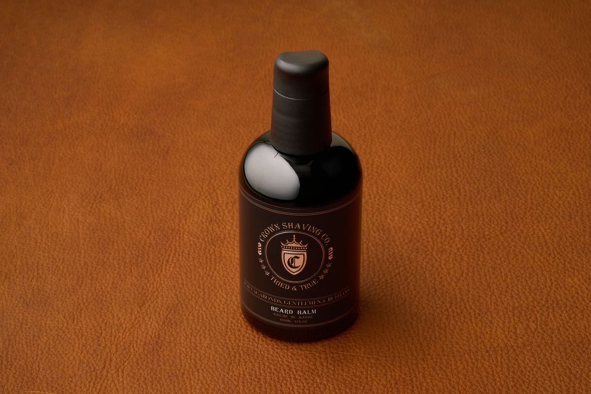 A bottle of Crown Shaving Co. Beard Balm, featuring a black label and cap, rests on a brown leather surface, providing a premium beard moisturizer for daily grooming.