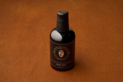 A bottle of Crown Shaving Co. Beard Balm, featuring a black label and cap, rests on a brown leather surface, providing a premium beard moisturizer for daily grooming.