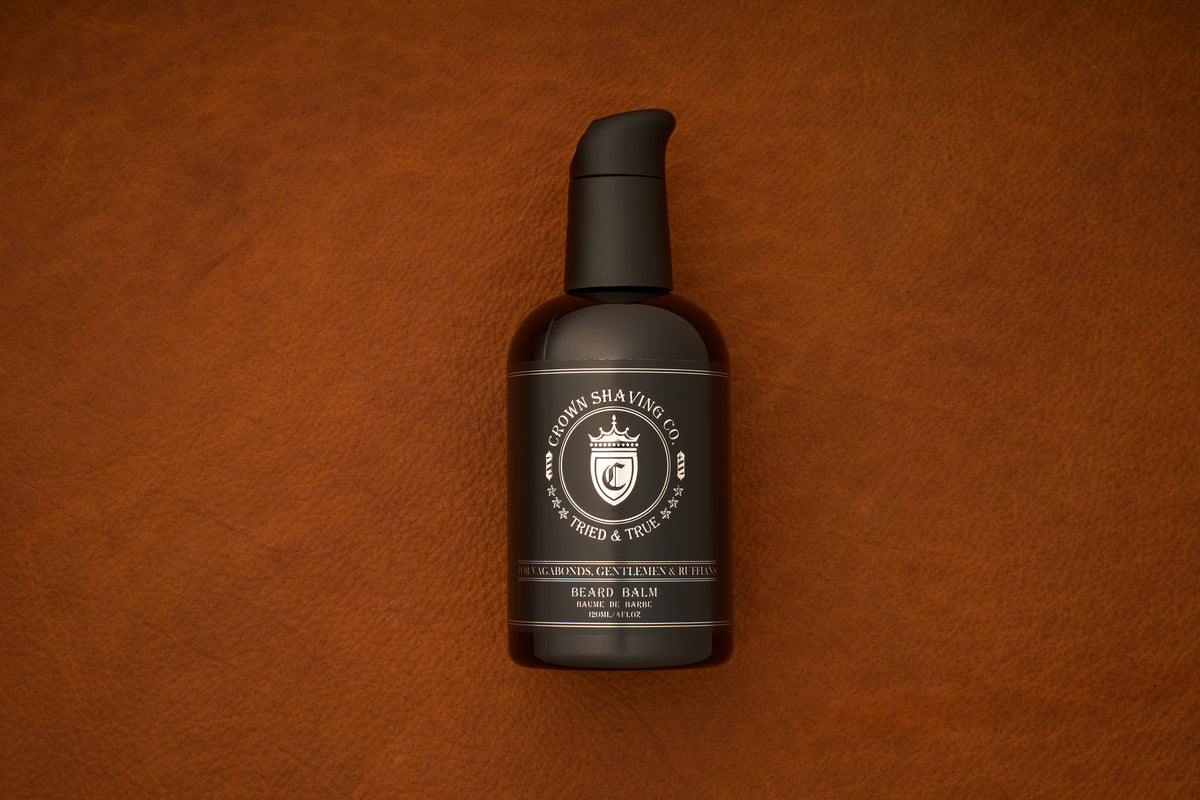 A black pump bottle of Crown Shaving Co. Beard Balm rests on a brown leather surface, featuring a white label with the crown logo and product information. This premium beard balm is ideal for daily grooming and care.