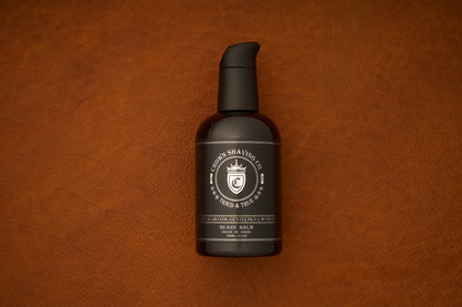 A black pump bottle of Crown Shaving Co. Beard Balm rests on a brown leather surface, featuring a white label with the crown logo and product information. This premium beard balm is ideal for daily grooming and care.