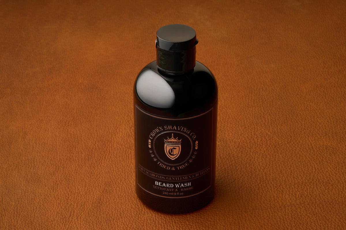 A 240mL black bottle of Crown Shaving Co. Beard Wash, a sulfate-free formula, rests on a brown leather surface. The label is adorned with gold accents and text, highlighting its commitment to premium beard care.