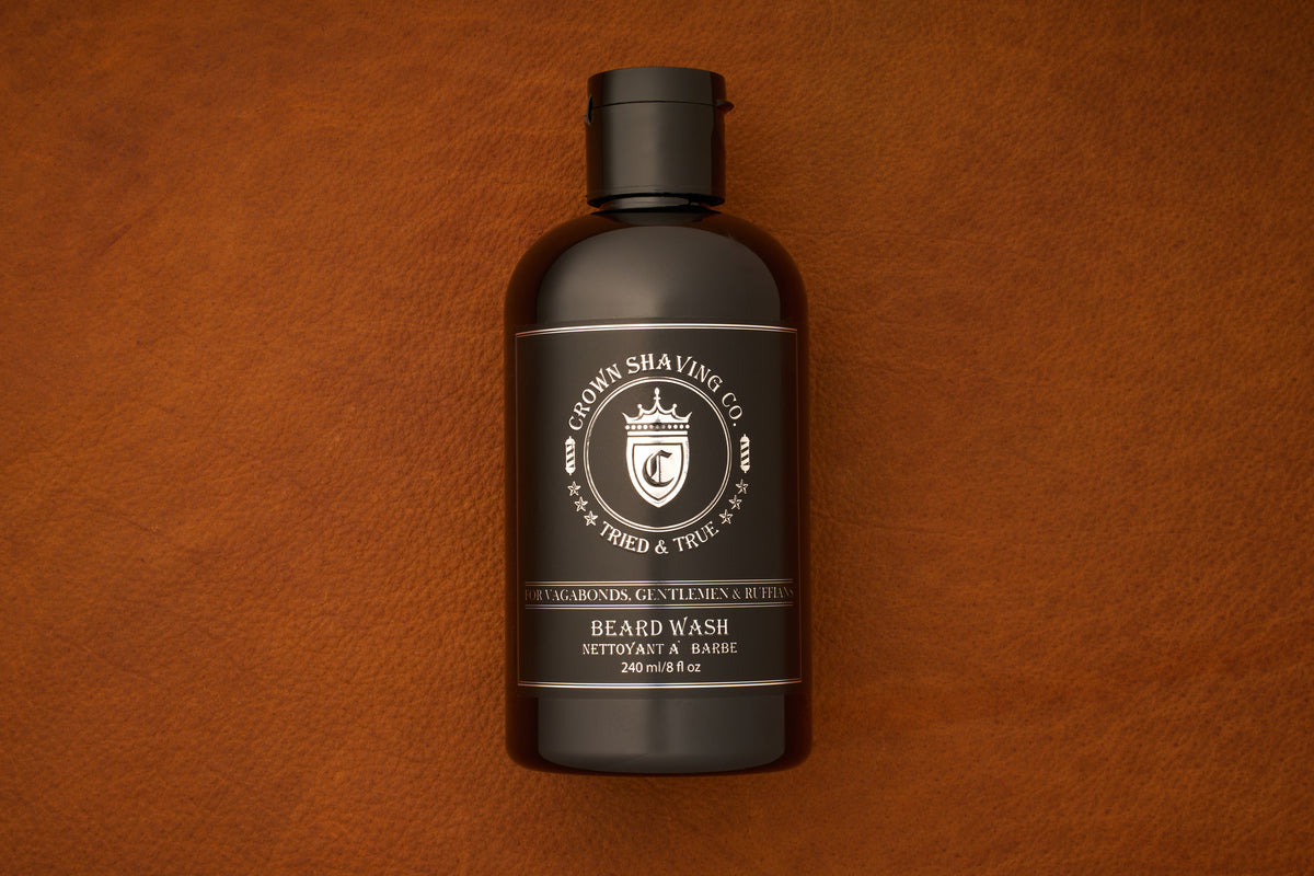 A 240mL bottle of Crown Shaving Co. Beard Wash rests on a brown leather surface. The dark brown bottle features a silver and white label with a crest, highlighting that it is a sulfate free product.