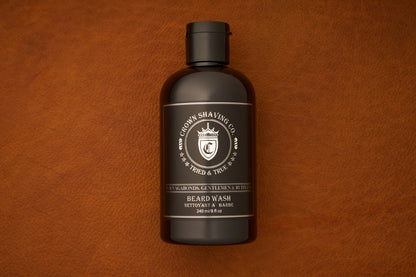 A 240mL bottle of Crown Shaving Co. Beard Wash rests on a brown leather surface. The dark brown bottle features a silver and white label with a crest, highlighting that it is a sulfate free product.