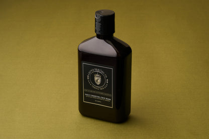 A dark bottle with a black cap and a gold and black label reading Crown Shaving Co. Daily Charcoal Face Wash stands on an olive green surface, highlighting its deep-cleansing face wash formula that harnesses the power of charcoal and tea tree to help prevent acne.