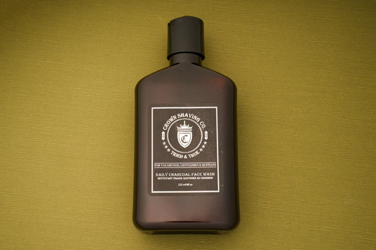 A brown bottle of Crown Shaving Co. Daily Charcoal Face Wash sits atop a green textured surface. Designed to help prevent acne for clear, refreshed skin, this deep-cleansing face wash is infused with charcoal and tea tree and features the signature crown logo alongside product details in white text.