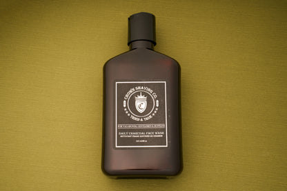 A brown bottle of Crown Shaving Co. Daily Charcoal Face Wash sits atop a green textured surface. Designed to help prevent acne for clear, refreshed skin, this deep-cleansing face wash is infused with charcoal and tea tree and features the signature crown logo alongside product details in white text.