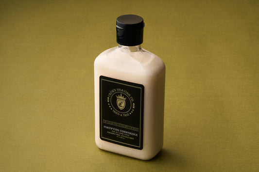 An 8 oz bottle of Crown Shaving Co. Fortifying Conditioner, featuring a black label and cap, rests on a green surface. The label displays the product name along with a circular logo crowned with text and highlights its sulfate free formula designed to keep hair healthy and nourished.