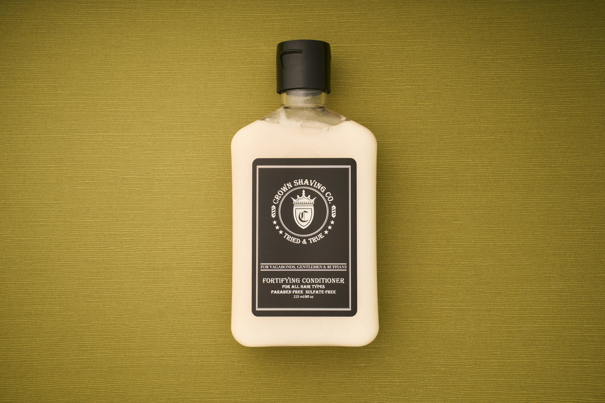 An 8 oz bottle of Crown Shaving Co. Fortifying Conditioner is placed on a textured olive-green surface. This sulfate-free, moisturizing conditioner comes in a rectangular bottle with a black cap and a black-and-white label.