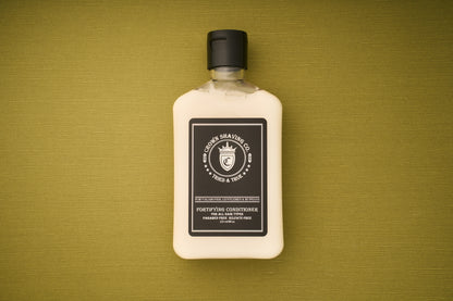 An 8 oz bottle of Crown Shaving Co. Fortifying Conditioner is placed on a textured olive-green surface. This sulfate-free, moisturizing conditioner comes in a rectangular bottle with a black cap and a black-and-white label.