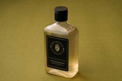 An 8oz bottle of Crown Shaving Co. Fortifying Shampoo, made of clear plastic with a black cap and label, rests on a green surface. The label is adorned with a gold crest and lettering that highlights its sulfate- and paraben-free formula. Visible bubbles inside the bottle emphasize its gentle yet effective scalp cleansing properties.