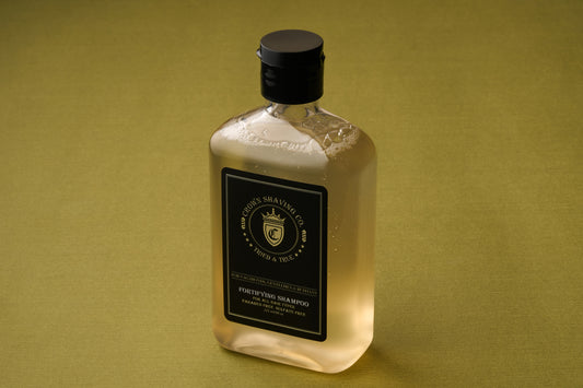 An 8oz bottle of Crown Shaving Co. Fortifying Shampoo, made of clear plastic with a black cap and label, rests on a green surface. The label is adorned with a gold crest and lettering that highlights its sulfate- and paraben-free formula. Visible bubbles inside the bottle emphasize its gentle yet effective scalp cleansing properties.