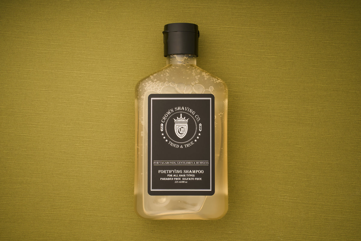 An 8oz bottle of Crown Shaving Co. Fortifying Shampoo, featuring a clear design with a black label and cap, rests on a textured olive-green background. The mostly full bottle, displaying some visible bubbles inside, emphasizes its sulfate- and paraben-free formula—perfect for gentle yet effective scalp cleansing.