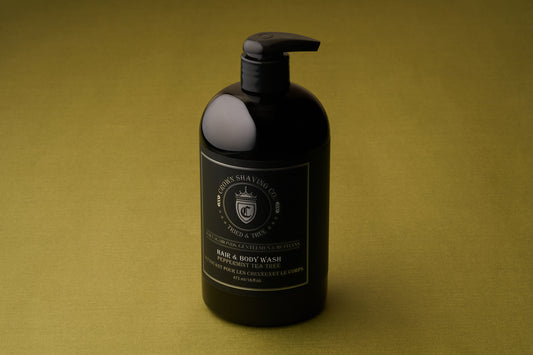 A black pump bottle labeled Crown Shaving Co. Hair and Body Wash – Peppermint & Tea Tree Oil stands on an olive green surface, with gold and white text on the front of the bottle highlighting that this is a sulfate- and paraben-free men’s body wash from Crown Shaving Co.