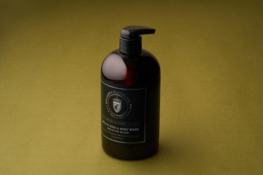A dark bottle with a pump dispenser containing Crown Shaving Co. Hair and Body Wash - Signature Scent 473mL/16oz—an invigorating men’s body wash that is sulfate and paraben free—sits on an olive-green surface, angled slightly to the right.