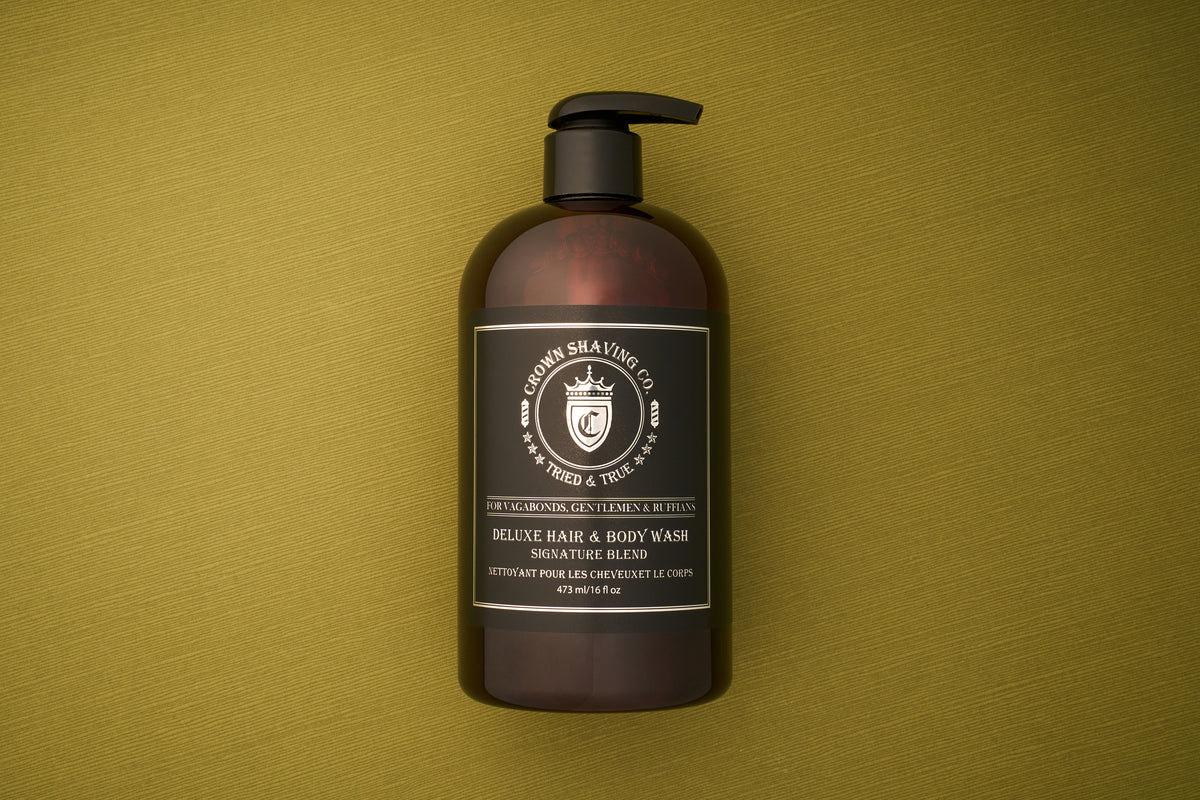 A brown pump bottle labeled Crown Shaving Co. Hair and Body Wash - Signature Scent 473mL/16oz rests on an olive green textured surface. The black label with white text features a crown logo and notes that this men’s body wash is sulfate and paraben free, offering a gentle yet effective cleanse.