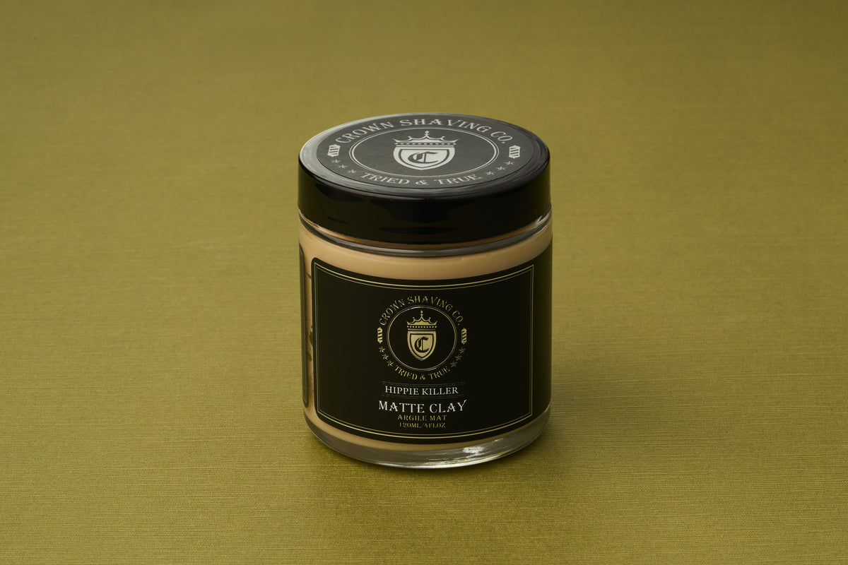 A glass jar of Crown Shaving Co. Hippie Killer Matte Styling Clay rests on a green surface. The jar, which contains a strong hold hair clay, showcases a black label and matching black lid adorned with gold and white text and graphics, offering a natural matte finish for your hairstyle.