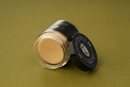 A glass jar of Crown Shaving Co. Hippie Killer Matte Styling Clay with a black lid displaying the logo lies on its side on an olive green surface. The open jar reveals the smooth beige strong hold hair clay inside, ideal for achieving a matte finish.