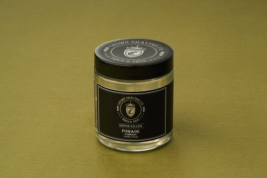 A clear jar of Crown Shaving Co. Hippie Killer Pomade, featuring a black label and lid, sits on an olive green surface. This water-soluble pomade is free from sulfates and parabens.