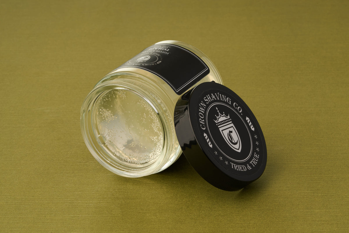 A glass jar of Crown Shaving Co. Hippie Killer Pomade lies on its side with the black lid off, resting on an olive green surface. The clear product inside, dotted with small bubbles, emphasizes its sulfate and paraben free formulation.