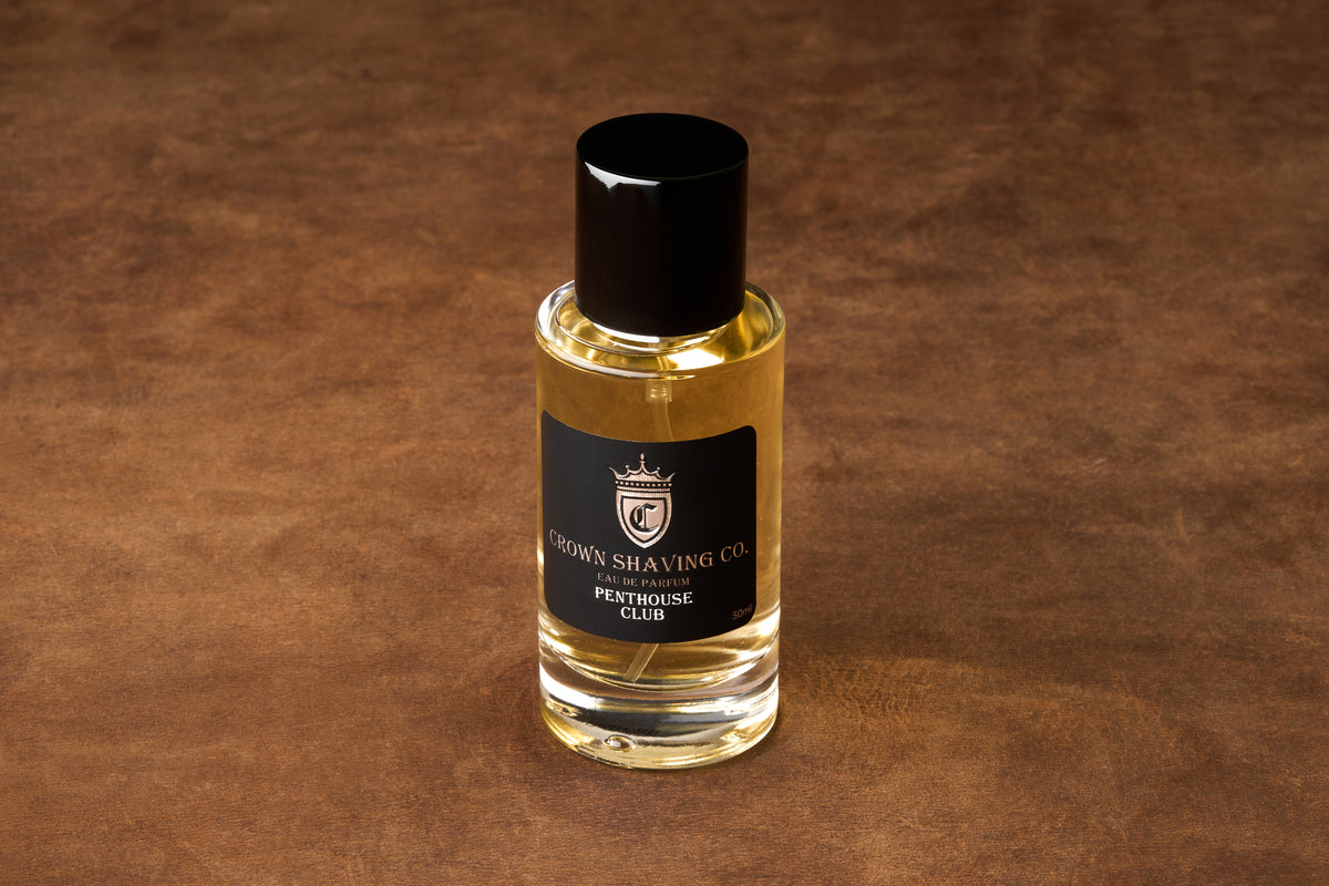 A bottle of Crown Shaving Co. Penthouse Club Eau de Parfum 50mL rests on a brown surface. Renowned for exceptional grooming, this fragrance comes in sleek packaging with a black label and cap, delivering a refined scent ideal for any sophisticated routine.