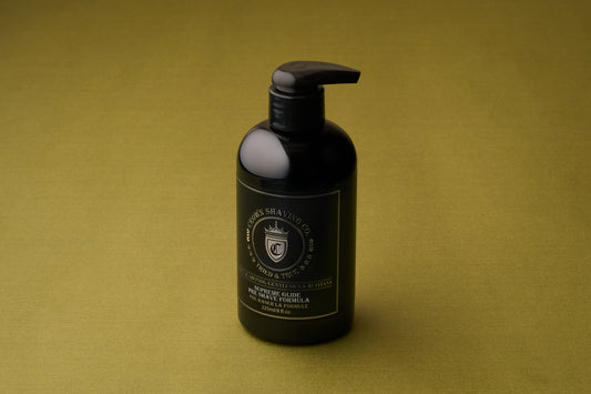 A black pump bottle of Crown Shaving Co. Preshave 225ml rests on a green surface, showcasing a crown logo along with gold and white text on the front. This pre-shave beard lubricant features a water-based formula designed to deliver a smooth and comfortable shaving experience.