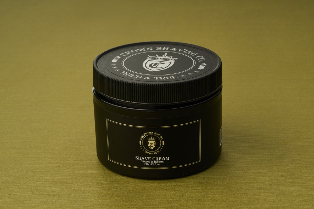 A black container of Crown Shaving Co. Shave Cream rests on an olive green surface, showcasing its logo and product label in white and gold. This peppermint shave cream is free from sulfates and parabens, providing a refreshing and gentle grooming experience.