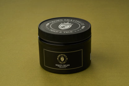 A black container of Crown Shaving Co. Shave Cream rests on an olive green surface, showcasing its logo and product label in white and gold. This peppermint shave cream is free from sulfates and parabens, providing a refreshing and gentle grooming experience.