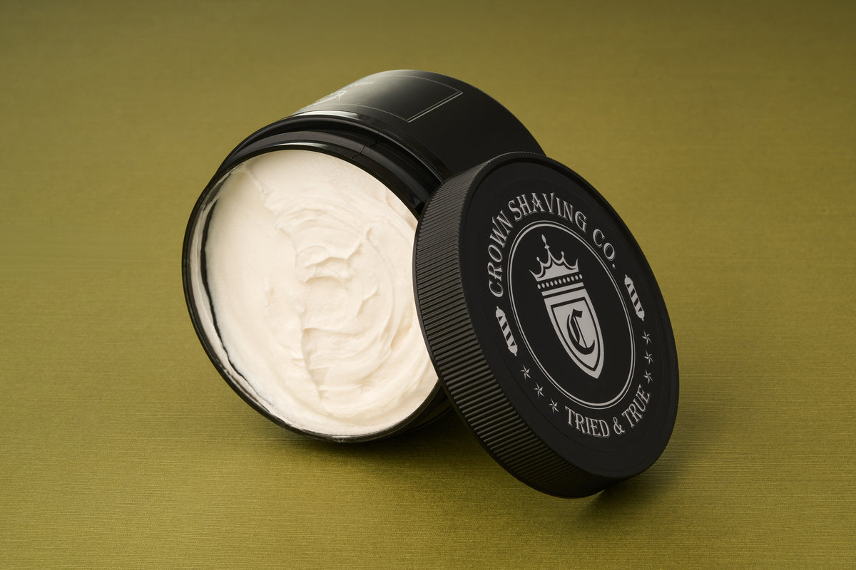 An open black container of Crown Shaving Co. Shave Cream rests on a green surface, its lid propped against the side to reveal the smooth, white cream inside. This luxurious peppermint formula is both sulfate-free and paraben-free, providing a refreshing and gentle shaving experience.