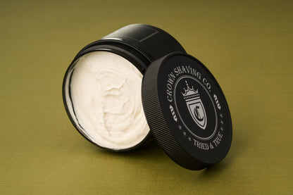 An open black container of Crown Shaving Co. Shave Cream rests on a green surface, its lid propped against the side to reveal the smooth, white cream inside. This luxurious peppermint formula is both sulfate-free and paraben-free, providing a refreshing and gentle shaving experience.