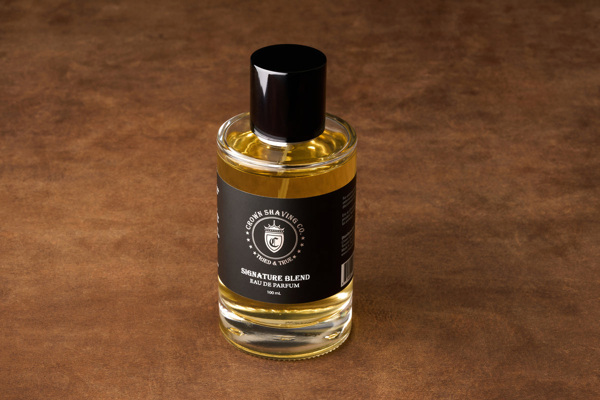A clear glass bottle of Crown Shaving Co. Eau de Parfum 100mL rests on a brown surface, featuring a black label and black cap—a refined addition to your grooming routine, with subtle Bay Rum notes that capture the classic Crown Shaving tradition.