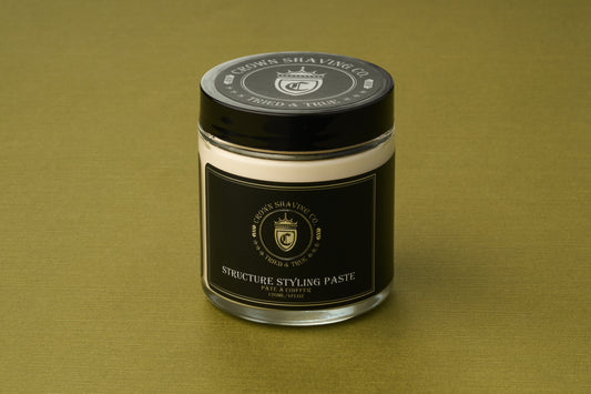 A glass jar of Crown Shaving Co. Structure Styling Paste sits on a green textured surface, featuring a black label with gold text and a gold crown logo on both the lid and front, emphasizing its premium matte finish and strong hold formula.