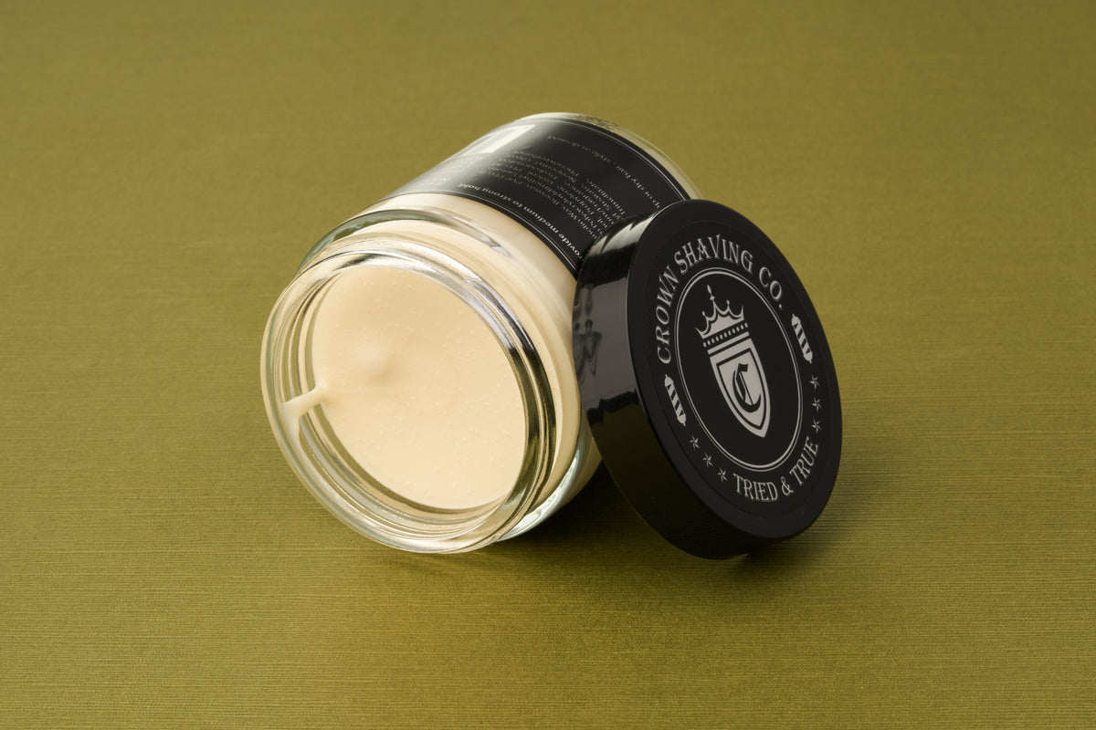A glass jar of Crown Shaving Co. Structure Styling Paste, featuring a cream-colored formula and topped with a black lid adorned with the white Crown Shaving Co. Trusted & True crest, sits on an olive-green surface—an ideal addition to your grooming routine, whether you’re using it as your go-to strong hold hair product for a flawless, matte finish.