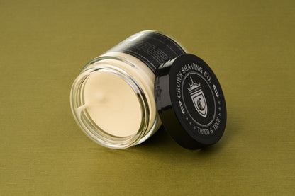 A glass jar of Crown Shaving Co. Structure Styling Paste, featuring a cream-colored formula and topped with a black lid adorned with the white Crown Shaving Co. Trusted & True crest, sits on an olive-green surface—an ideal addition to your grooming routine, whether you’re using it as your go-to strong hold hair product for a flawless, matte finish.