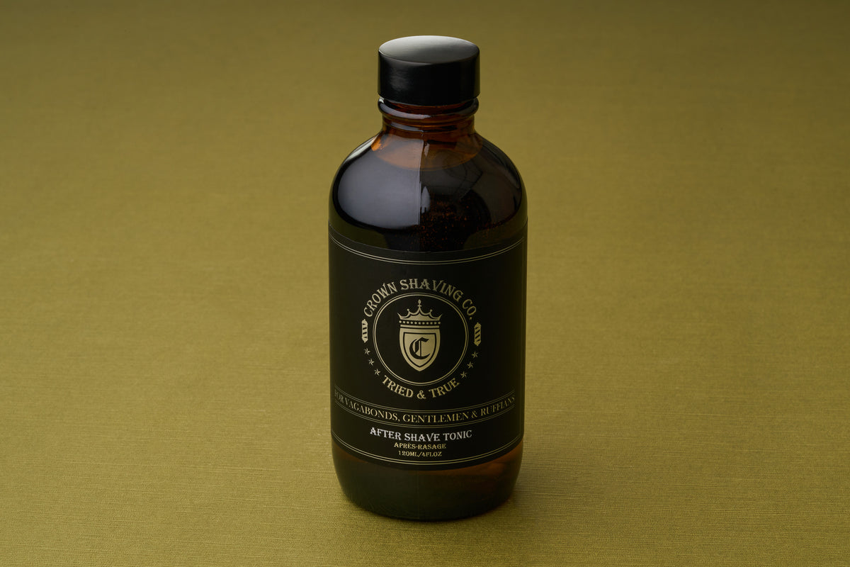 A brown glass bottle of Crown Shaving Co. Aftershave Tonic, featuring a black label with gold lettering, rests on a simple olive-green surface.