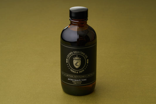 A brown glass bottle of Crown Shaving Co. Aftershave Tonic, featuring a black label with gold lettering, rests on a simple olive-green surface.