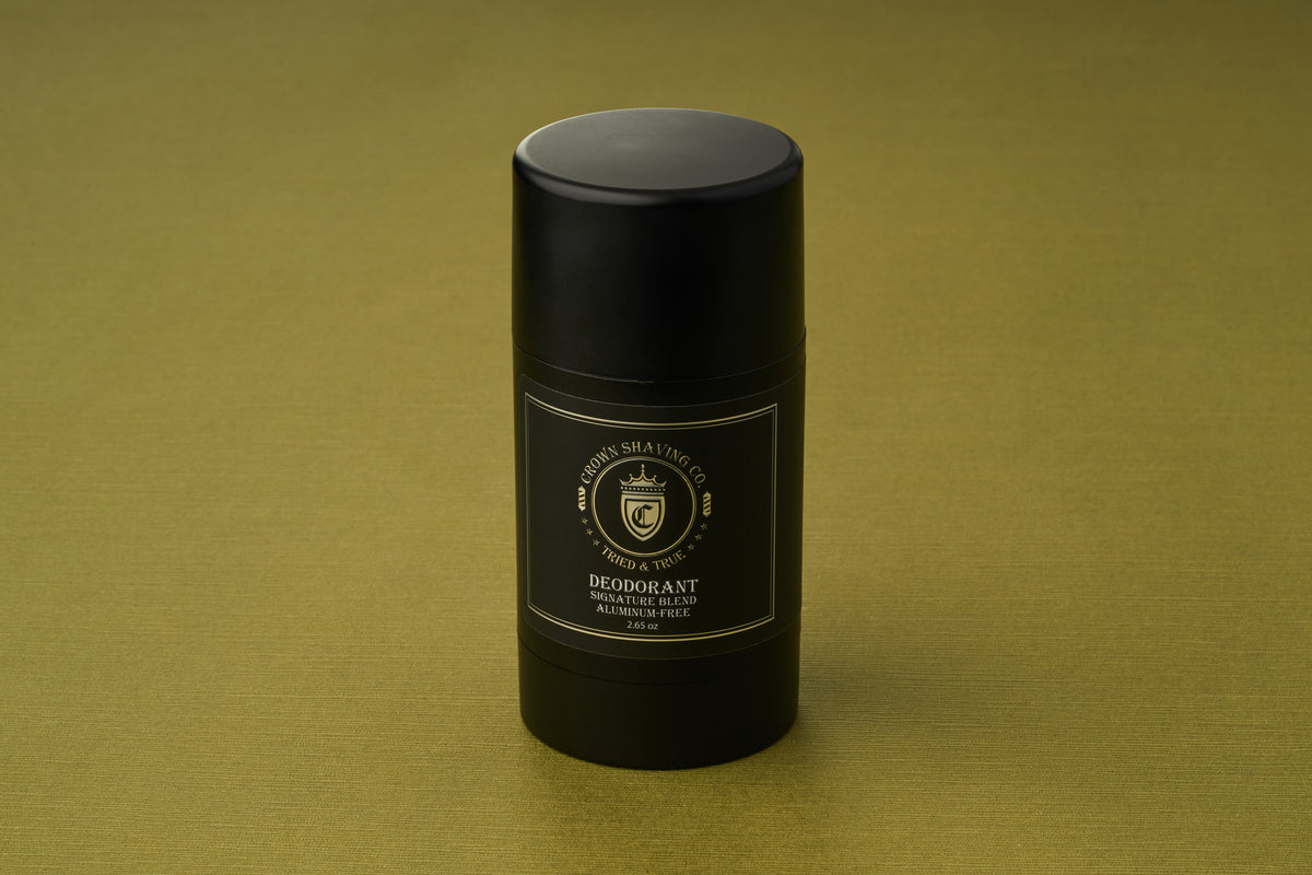 A black cylindrical stick of Crown Shaving Co. Aluminum-Free Deodorant, featuring a gold and white label, stands upright on an olive green textured surface. This deodorant provides reliable, all-day protection without aluminum.