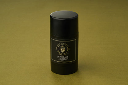 A black cylindrical stick of Crown Shaving Co. Aluminum-Free Deodorant, featuring a gold and white label, stands upright on an olive green textured surface. This deodorant provides reliable, all-day protection without aluminum.