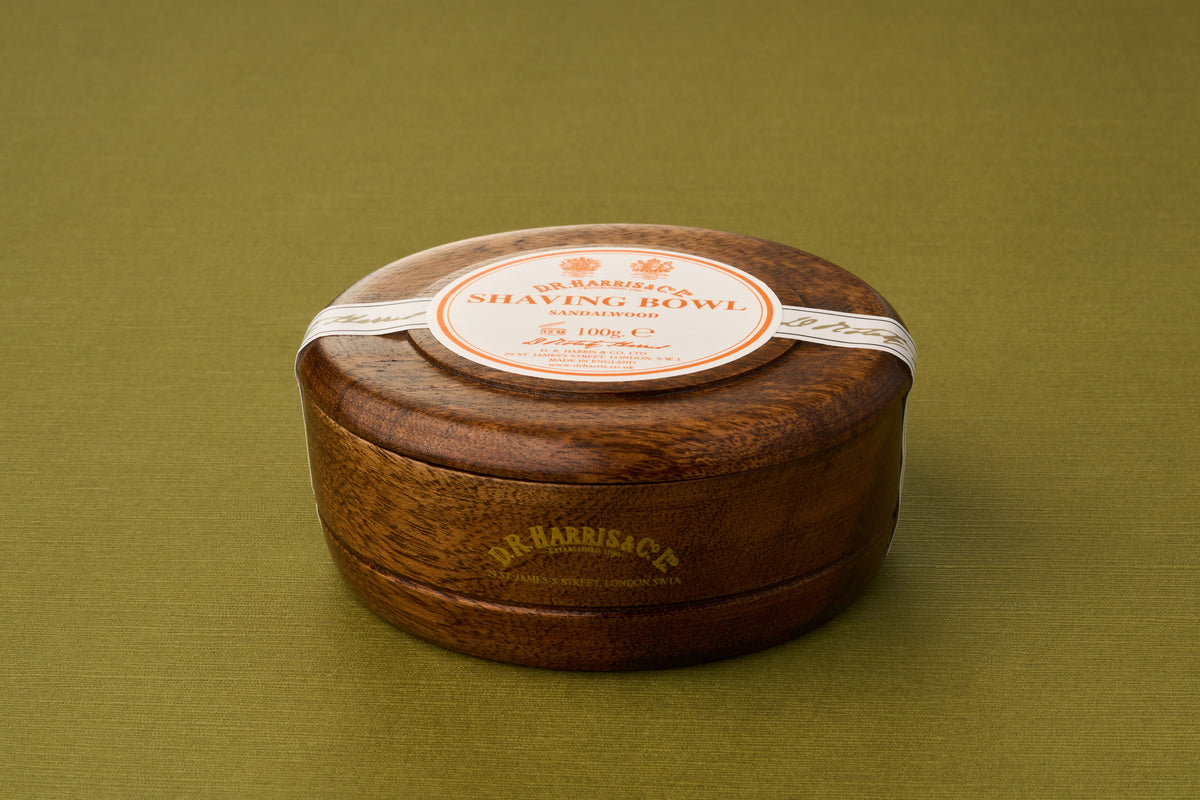 A round, closed wooden shaving bowl featuring a white and orange D.R. Harris & Co. label rests on an olive green surface, containing the D.R. Harris Sandalwood Shaving Soap—a classic option for those seeking a luxurious, zero-waste shaving routine.