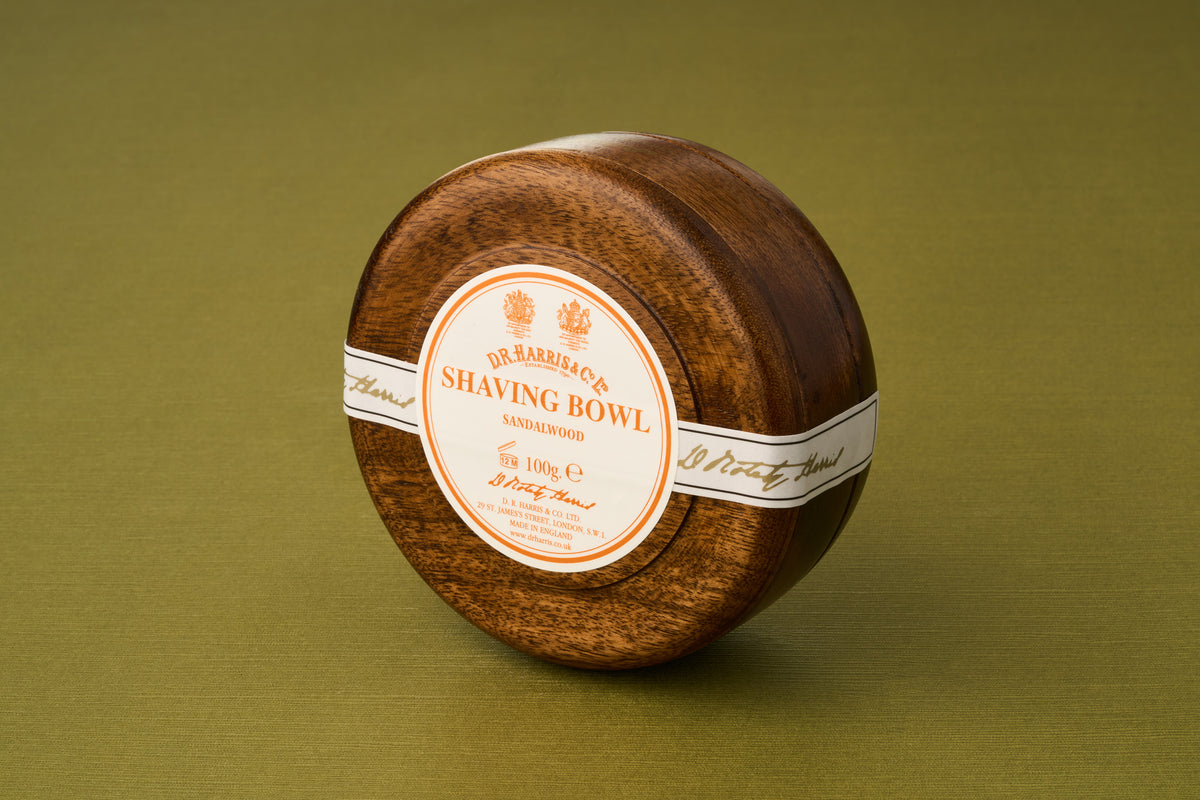 A round wooden bowl containing D.R. Harris Sandalwood Shaving Soap, distinguished by its white and orange label, rests on a green surface—offering a luxurious shaving soap experience that enhances your grooming routine while supporting zero-waste shaving.
