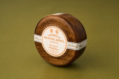 A round wooden bowl containing D.R. Harris Sandalwood Shaving Soap, distinguished by its white and orange label, rests on a green surface—offering a luxurious shaving soap experience that enhances your grooming routine while supporting zero-waste shaving.