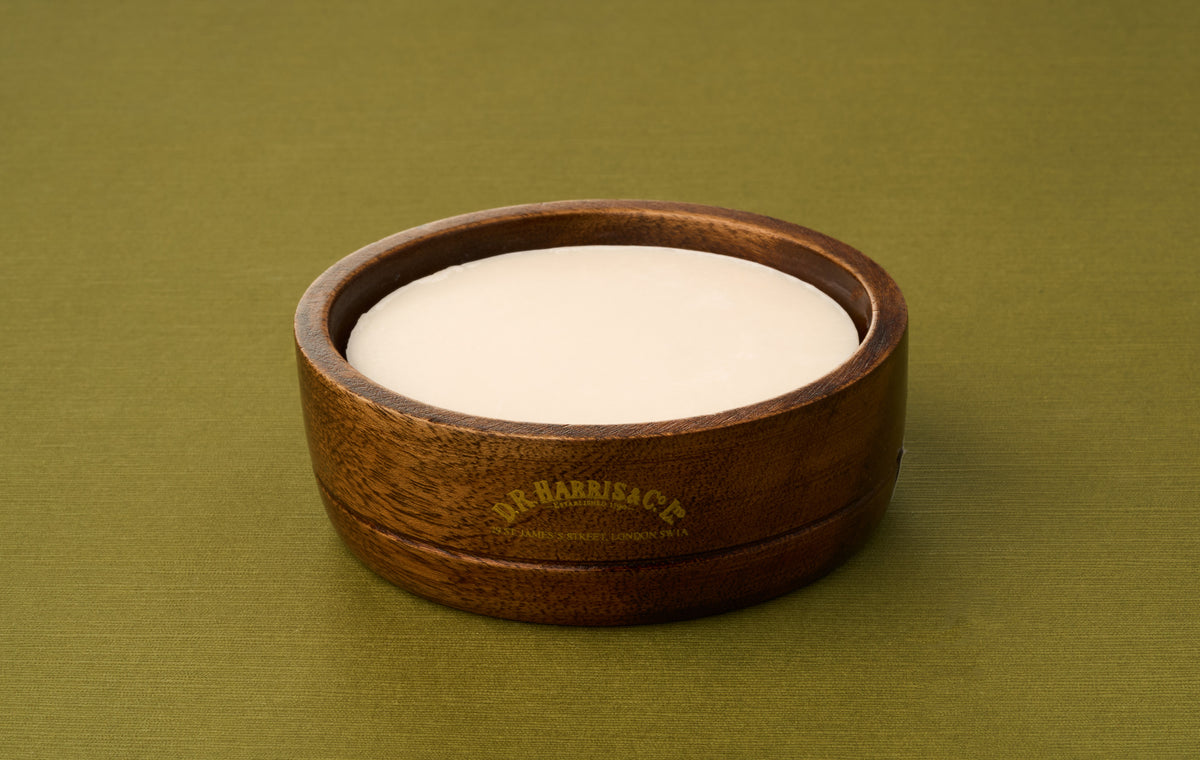 A round bar of D.R. Harris Sandalwood Shaving Soap rests in a brown wooden bowl labeled D.R. Harris & Co., set atop a green textured surface to offer an elegant, zero-waste shaving experience.
