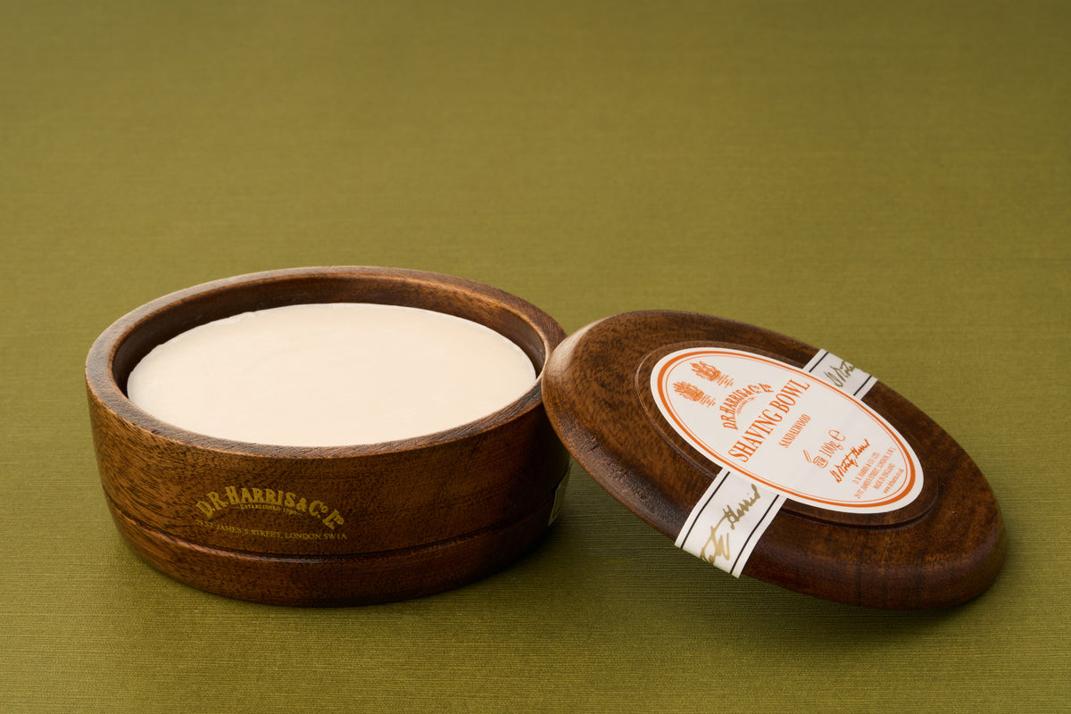 A round wooden container of D.R. Harris Sandalwood Shaving Soap sits on a green surface with its lid slightly ajar to reveal the soap inside. The lid features an orange label with text, making this an elegant zero-waste option for discerning shavers.