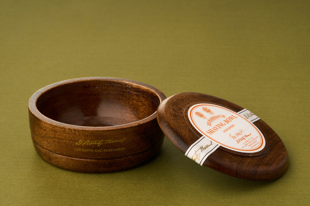 A D.R. Harris Sandalwood Shaving Soap wooden bowl sits on a green surface, its polished interior empty and its matching lid—adorned with a distinctive white and orange label—resting nearby: an elegant, zero-waste option for the eco-conscious shaving enthusiast.