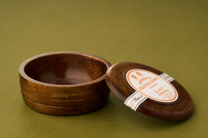 A D.R. Harris Sandalwood Shaving Soap wooden bowl sits on a green surface, its polished interior empty and its matching lid—adorned with a distinctive white and orange label—resting nearby: an elegant, zero-waste option for the eco-conscious shaving enthusiast.