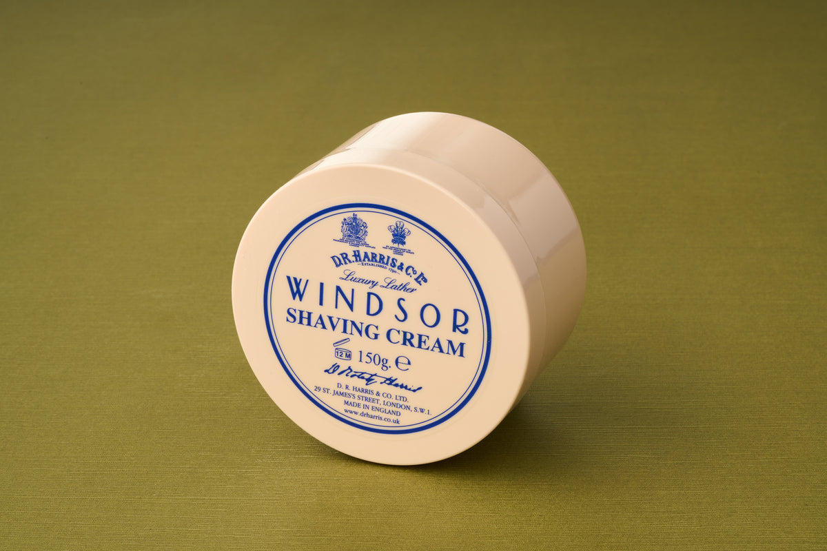 A beige, round container of D.R. Harris Windsor Shaving Cream, a luxury shaving product from the brand, sits on a green surface. The lid displays blue text and details with the brand name and product information.