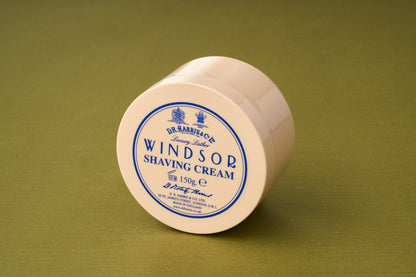 A beige, round container of D.R. Harris Windsor Shaving Cream, a luxury shaving product from the brand, sits on a green surface. The lid displays blue text and details with the brand name and product information.