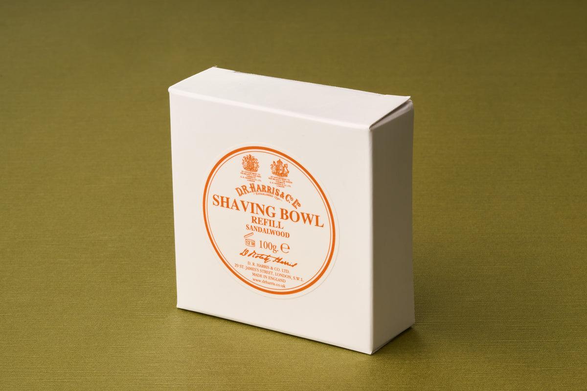 A white box labeled D.R. Harris Sandalwood Shaving Soap rests on a green surface. The label is circular, printed in orange with decorative text, and displays a weight of 100g. This luxurious shaving soap pairs classic sandalwood fragrance with sustainable, zero-waste grooming for an effortless shave.